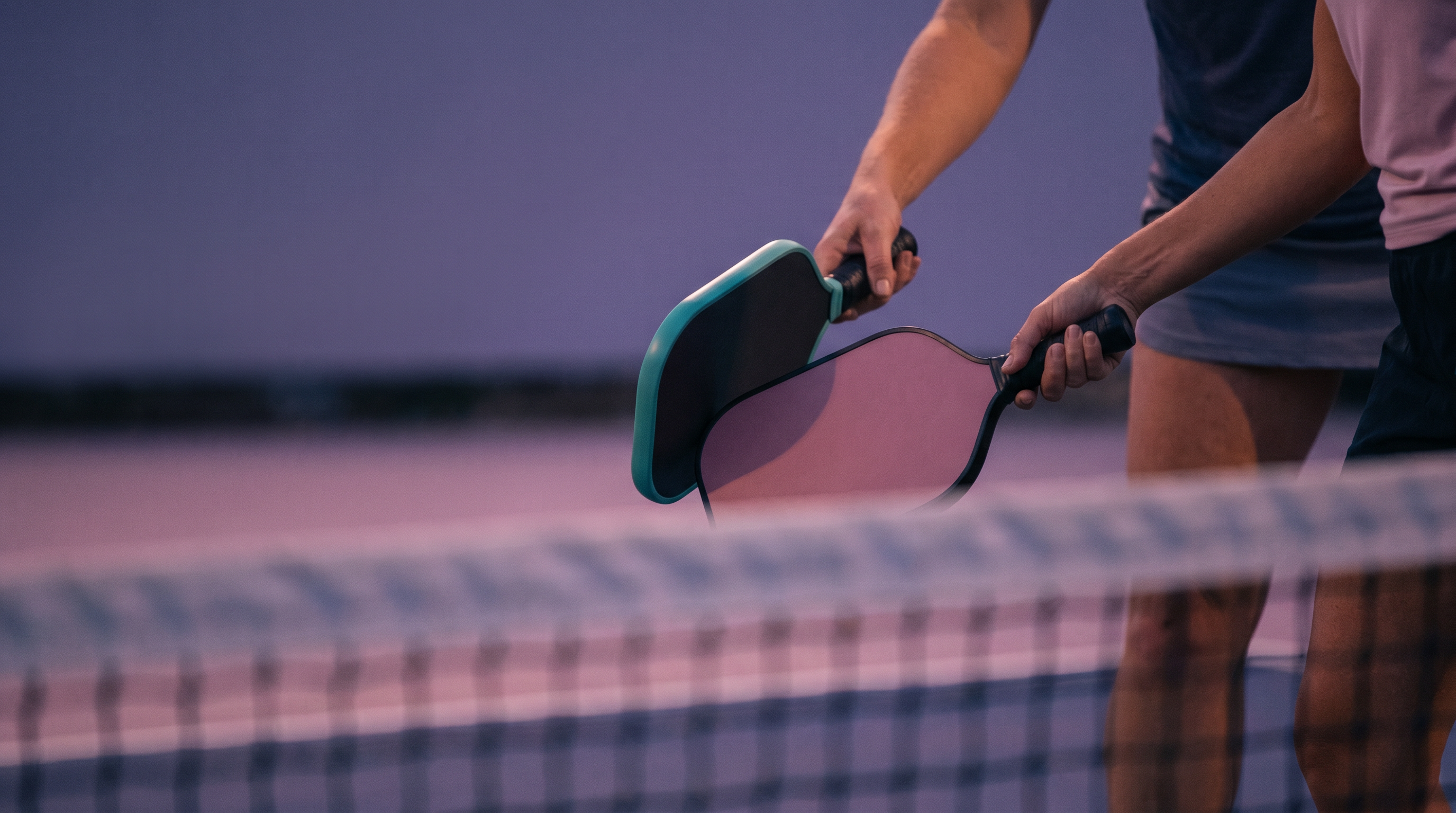 Close-up of two pickleball paddles tapped together at the net in the "good game" gesture, dusk light, no faces visible.