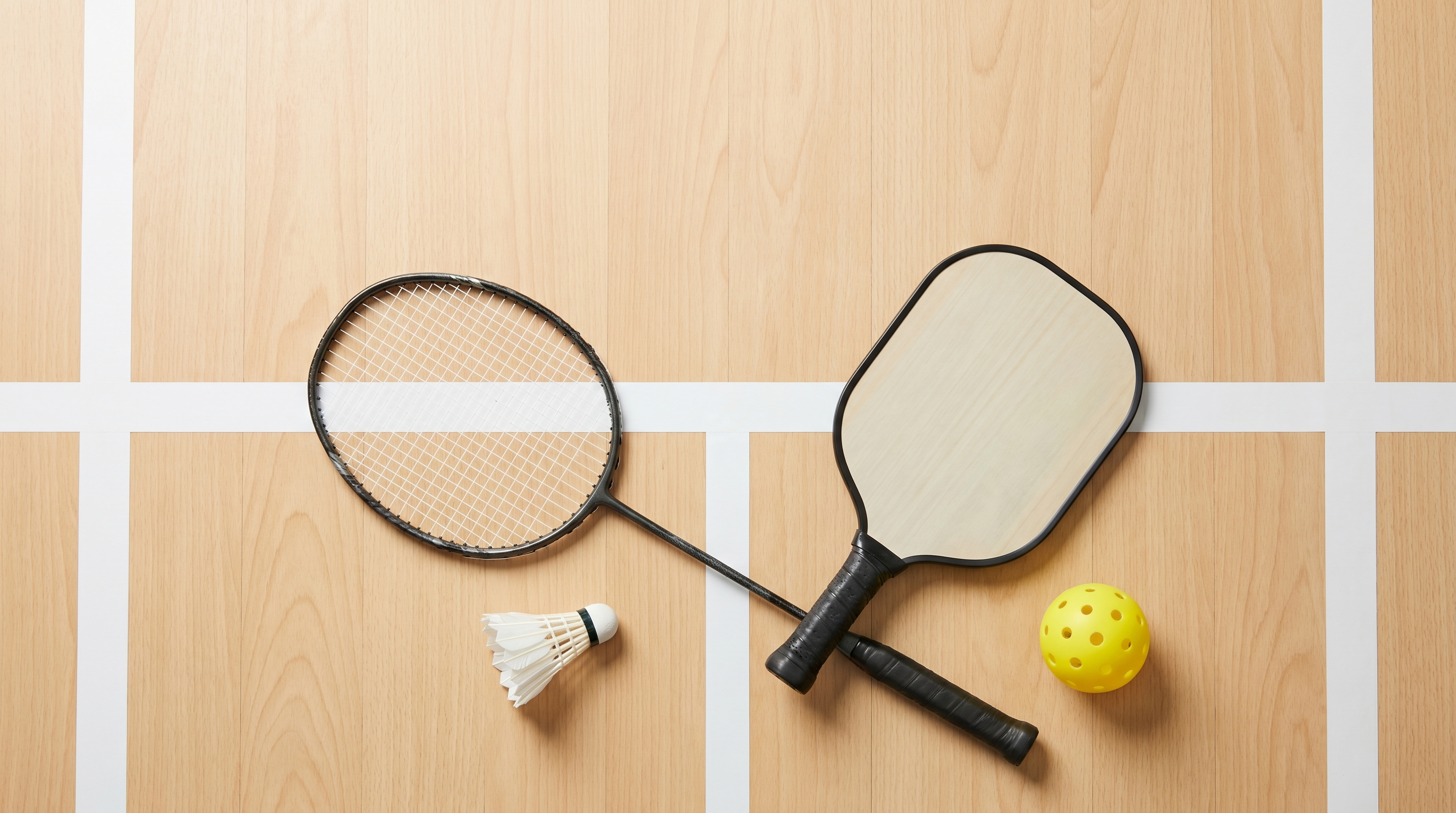 Top-down composition of a badminton racquet and pickleball paddle crossed on deep maroon velvet, shuttlecock and pickleball alongside.